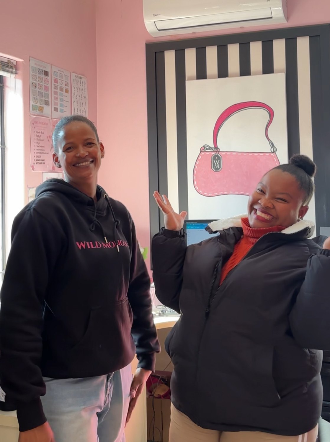 Denina and Lorikah laughing together in the Wild Mongoo shop in George beside the pink handbag artwork