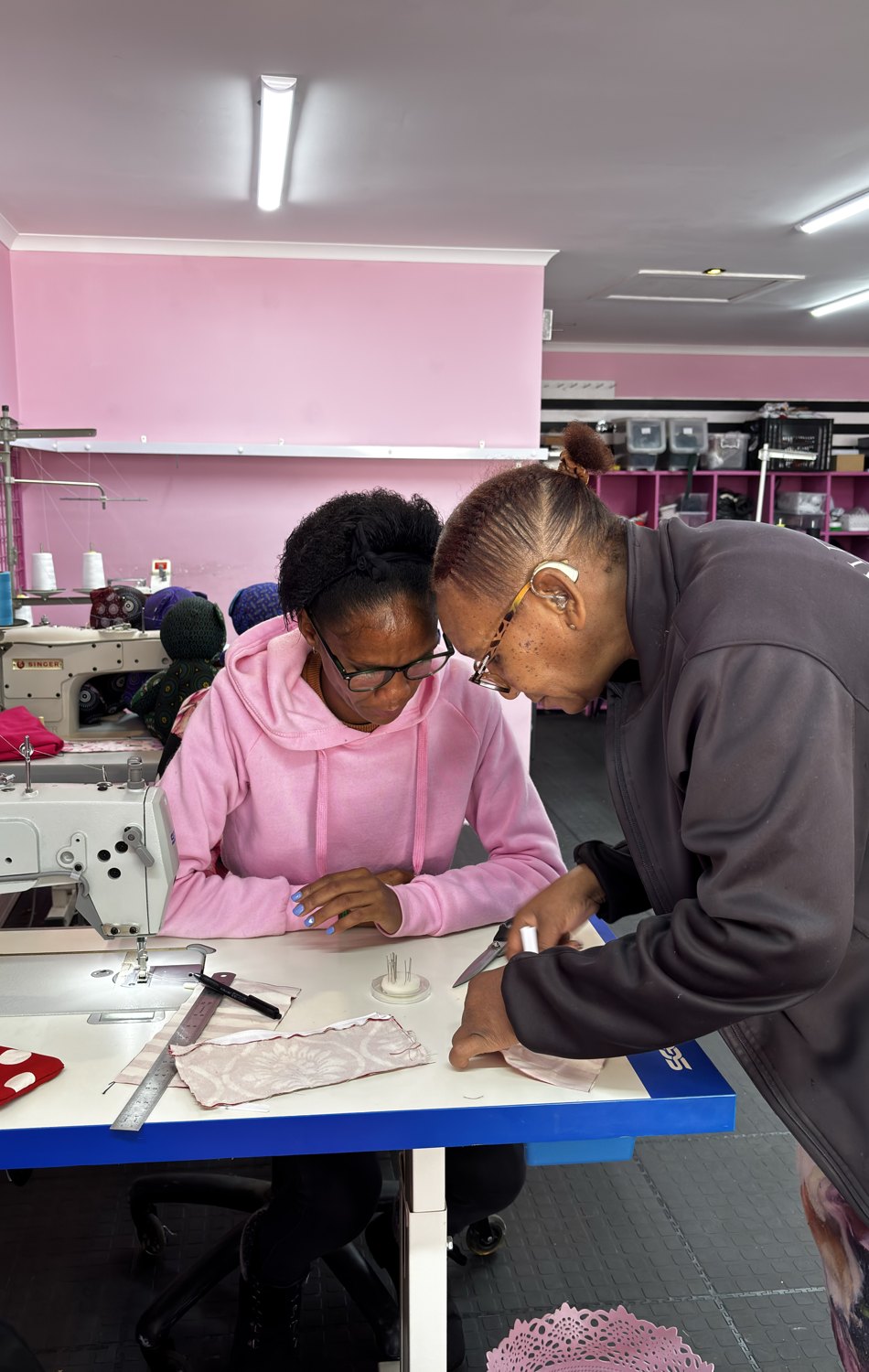 Betty teaching Ricky at a sewing machine in the Wild Mongoo workshop, sharing skills handmade in George