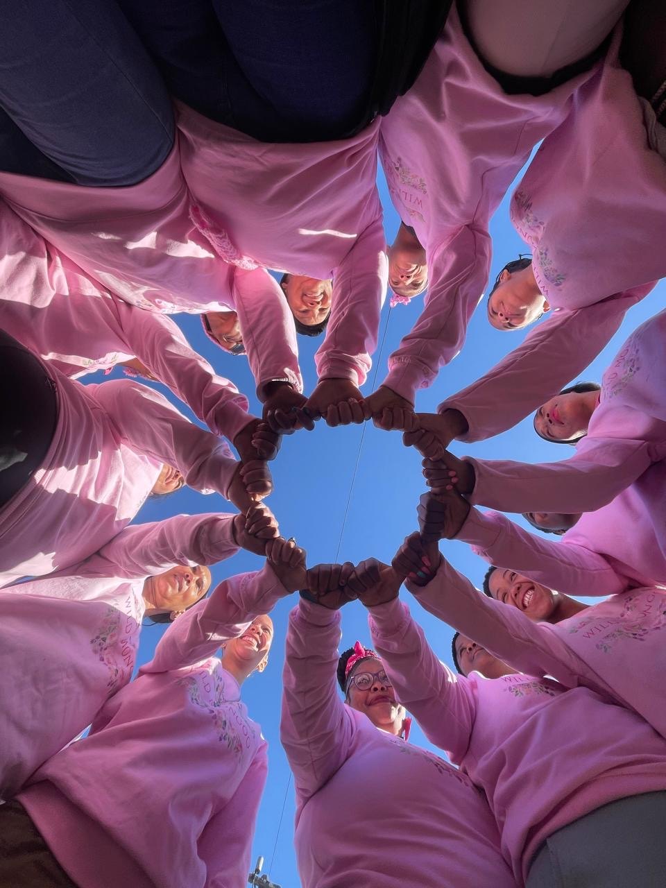 The Wild Mongoo women in a circle photographed from below against a blue sky, hands joined in the centre
